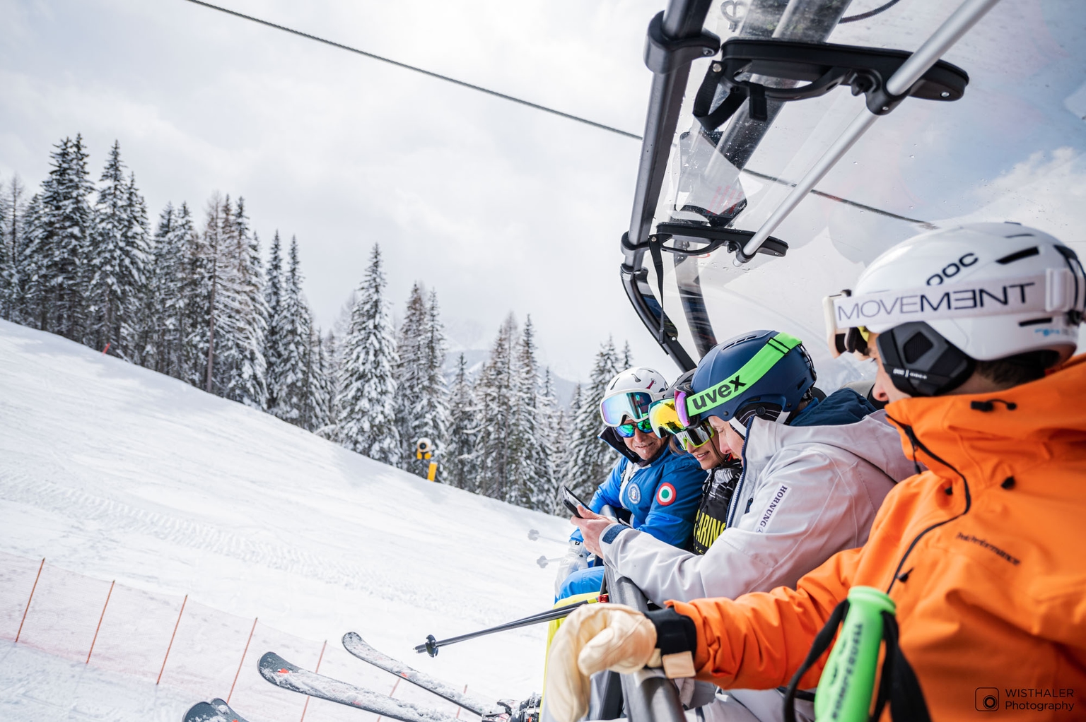 Drei Skifahrer*innen sitzen in einem Sessellift und schauen gemeinsam mit ihrem Skilehrer in ein iPad.