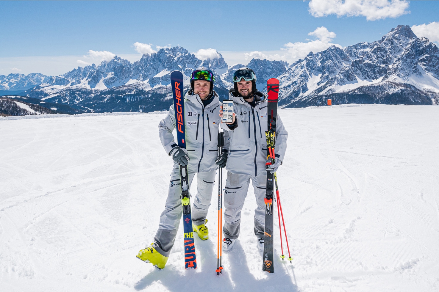 Zwei Ski-Ausbilder stehen in grauen Skiklamotten vor einem schönen Bergpanorama im Schnee. Ihre Ski lehnen an ihren Schultern. Die rechte Person hält ihren Handyscreen zur Kamera. Auf dem Screen ist die How2ski App zu sehen.