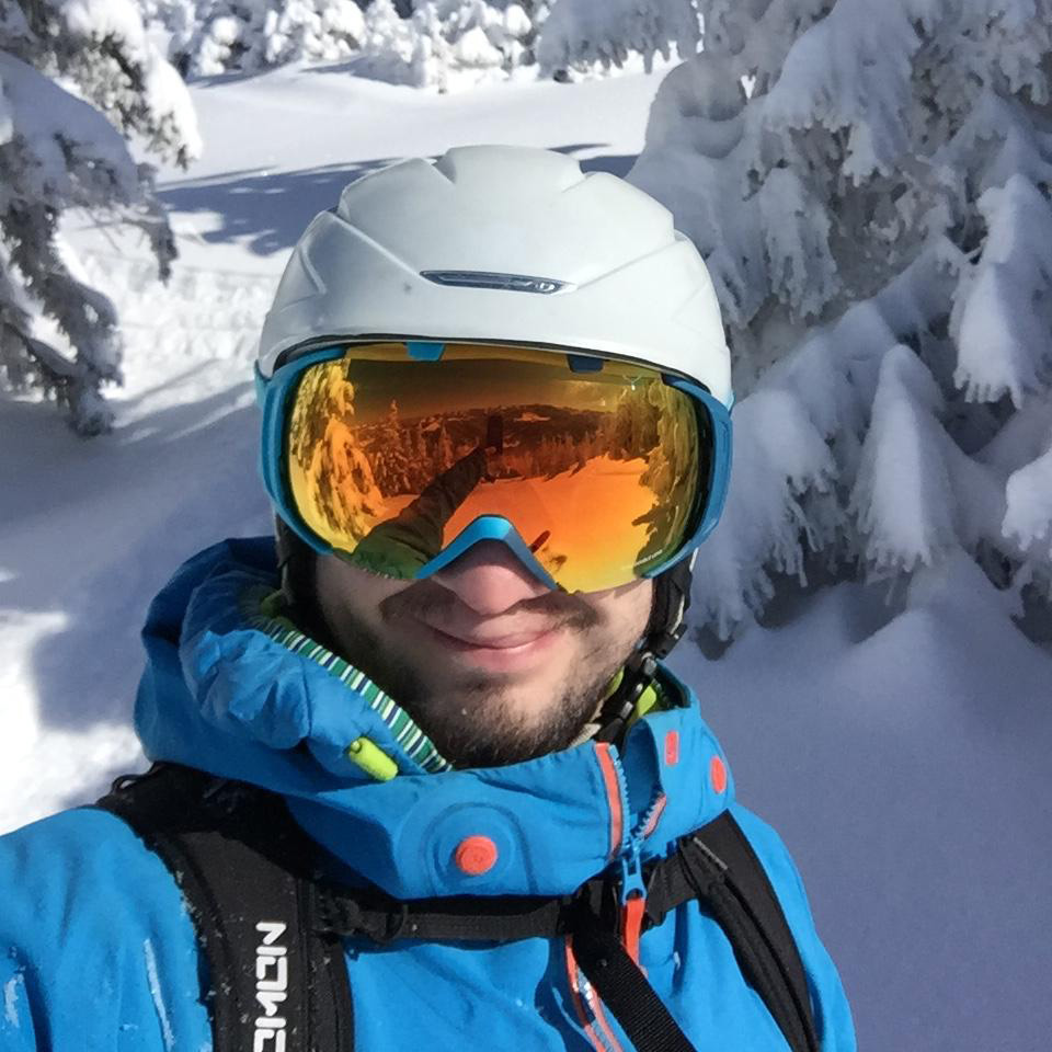 Junger Mann mit Bart, Skihelm und orange verspiegelter Brille grinst in die Kamera. Er trägt blaue Skiklamotten und einen Rucksack. Im Hintergrund sind schneebedeckte Bäume zu sehen.