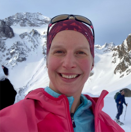 Katha Weber steht grinsend vor einem schneebedeckten Berg. Sie trägt ein lila Stirnband und eine pinke Weste. Die Sonnebrille hat sie auf dem Kopf und im Hintergrund ist ein Skitourengänger zu sehen.